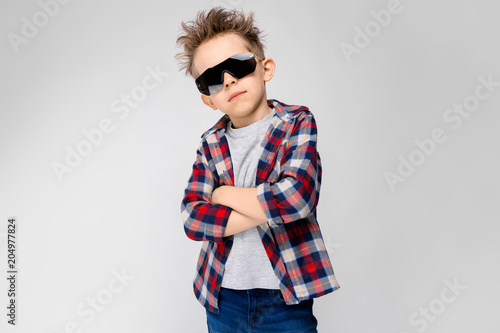 A handsome boy in a plaid shirt, gray shirt and jeans stands on a gray background. The boy in the black sunglasses. The boy folded his arms over his chest