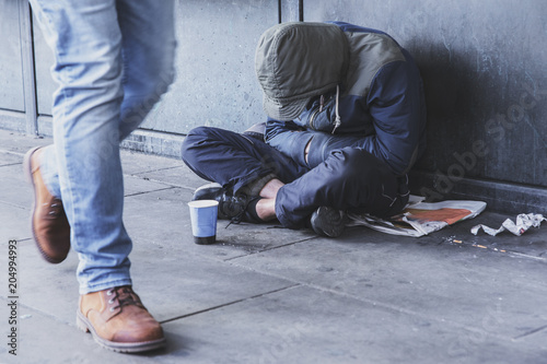 Homeless man sitting on the street in the shadow of the building and begging for help and money. Problems of big modern cities.