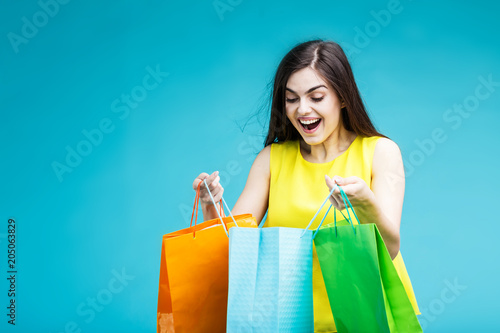 Happy smiling brunette girl wears yellow shirt holding colorful shopping bags on blue background, shopaholic sale concept