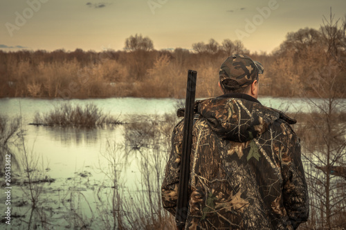 Hunter man shotgun camouflage exploring flood river hunting season rear view sunset
