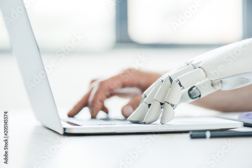 selective focus of human and robot hands typing on laptop at workplace