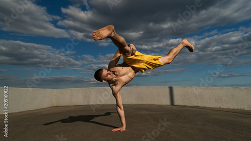 Tricking on street. Martial arts. Man performs blow with support of his hand barefoot. Shooted from bottom foreshortening against sky.