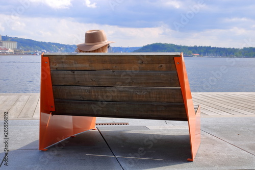 Drewniana ławka z czerwonymi metalowymi bokami, od tyłu, na betonowo-drewnianym nabrzeżu, jakby molo, na niej siedzi turysta w słomkowym kapeluszu i ciemnych okularach, patrzy na zatokę i drugi brzeg
