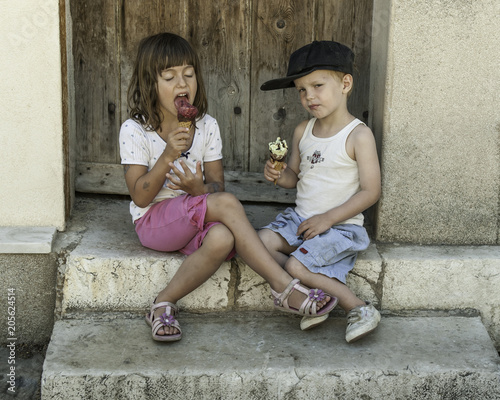 Portrait of two untidy scruffy dirty young caucasian french children blissfully eating ice-cream in front of rustic doorway in summer