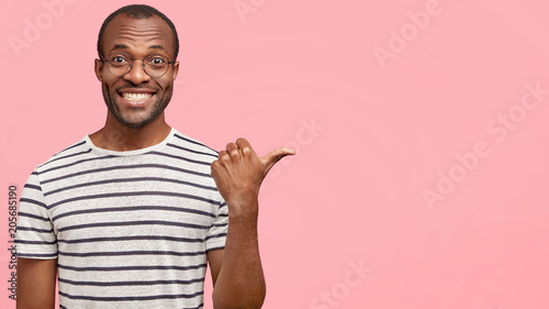 Happy dark skinned African male with pleasant appearance, indicates with thumb aside at pink background, attracts audience attention while presents well done project on meeting, has shining smile