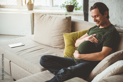 Dreaming of future child. Cheerful young man is sitting on sofa as if he is pregnant. He is touching his belly with love and smiling. Copy space 