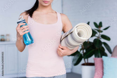 cropped shot of young woman with rolled yoga mat and bottle of water at home