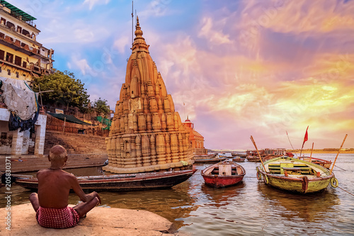 Old man meditates at Varanasi India at the Ganges river bank with ancient architecture and boats at sunrise.