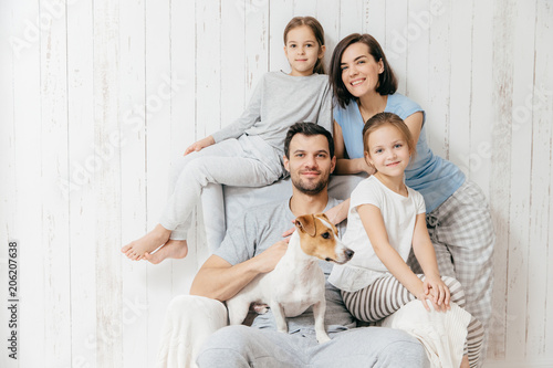 Family portrait. Happy parents with their two daughters and dog pose together against white background, spend free time at home, being in good mood. Mother, father and small sisters pose indoor