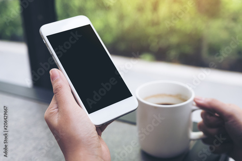Mockup image of hands holding white mobile phone with blank black desktop screen while drinking coffee in cafe