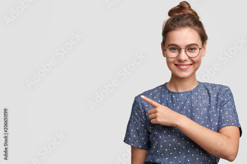 Use this copy space wisely. Cheerful Caucasian female with glad expression, indicates left with positive expression, dressed in elegant clothes, happy to advertise something on blank white wall