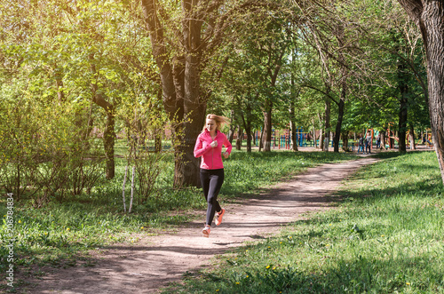 A young athletic girl in a pink and black sports suit jogging along the path in the park in the morning.
