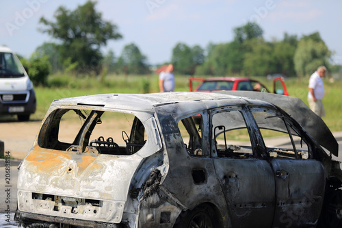 Spalony samochód osobowy na ulicy i poszkodowani ludzie.