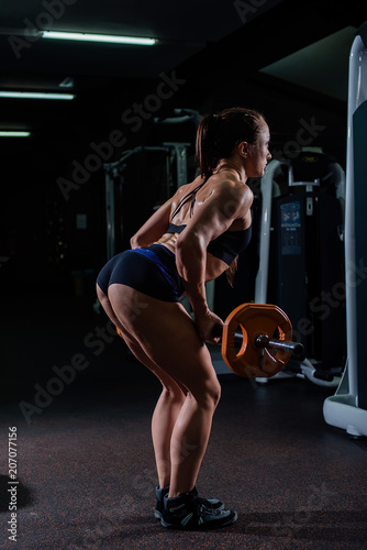 A strong, muscular, girl in black shorts, performs an exercise of deadlift, with a sports bar. Dark gym.