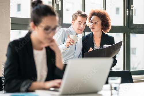 Two young malicious employees gossiping about their hard-working colleague in the office