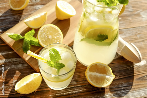 Iced lemonade pitcher, wooden juicer & one glass of cold citrus beverage with lemon slices, mint leaves & yellow straw on brown grunged wood textured table sunny day, background. Close up, copy space.