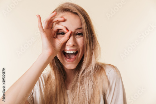 Image of amusing caucasian woman wearing casual clothing laughing and looking at you through hole made by fingers like ok sign, isolated over beige background