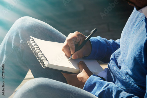 Attractive casually dressed young american african man making notes in copybook pad. Student preparing for lesson at college