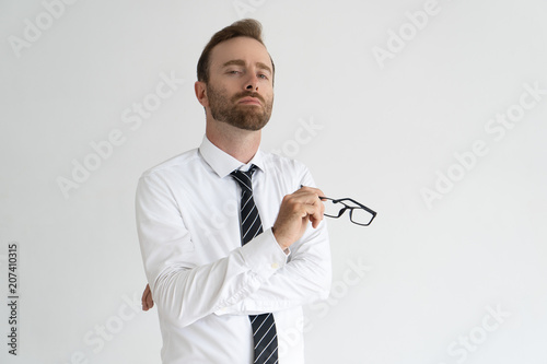 Young arrogant business expert posing for camera. Vain serious Caucasian man with pained face expression holding glasses. Self importance and ego concept