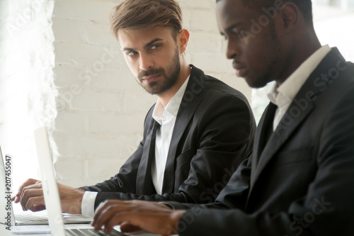 Suspicious cunning male Caucasian worker looking at serious working African American colleague, feeling mad and sneaky distrusting, having doubts, planning. Concept of office relationships, jealousy