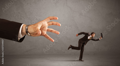 Young miniature businessman running from a big hand with grey background