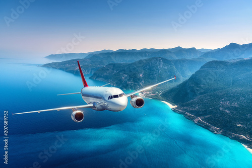 Airplane is flying over islands and sea at sunrise in summer. Landscape with white passenger airplane, seashore, mountains, sky, and blue water. White passenger aircraft. Travel and resort. Tourism