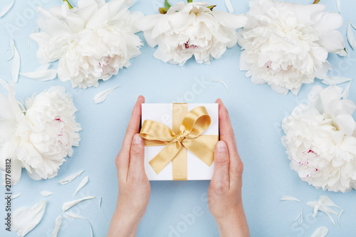 Womans hands giving gift or present box decorated white peony flowers on pastel table top view. Flat lay composition for birthday or wedding.