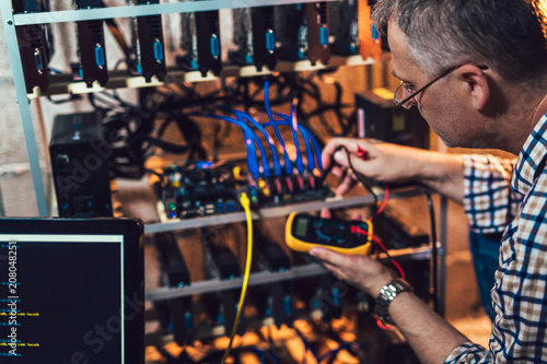 Programmer holding a wattmeter measuring cryptocurrency mining rig electrical energy consumption.