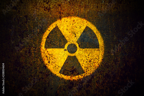 Round yellow radioactive (ionizing radiation) danger symbol painted on a massive rusty metal wall with dark rustic grungy texture background with vignetting.