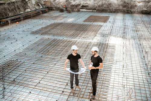 Two builders in t-shirts and protective helmets standing with drawings on the concrete foundation at the construction site