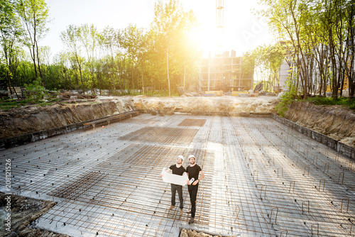 Wide angle view on the concrete foundation at the construction site with two builders standing with drawings during the sunset