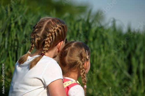 Dwie dziewczynki, blondynki uczesane w warkoczyki, nastolatka i kilkulatka, siedzą razem, blisko siebie, na zewnątrz, tyłem, w tle bujna wiosenna (letnia) zieleń