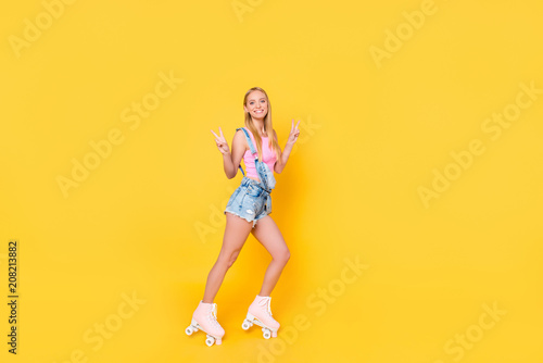 Full size body portrait of attractive pretty girl riding on roller skates gesturing v-signs with two hands looking at camera isolated on yellow background