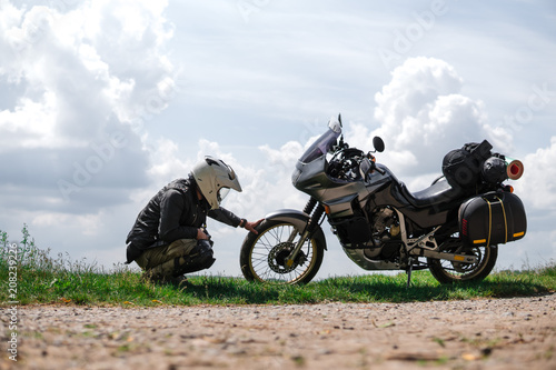 Rider Man and off road adventure motorcycles with side bags and equipment for long road trip, river and clouds on background, problems on the road in travel, service and punched wheel, faulty concept