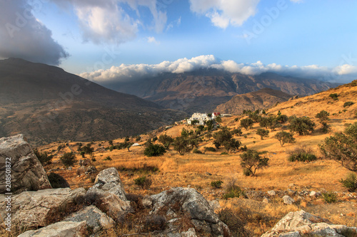 Ausblick über Kreta mit Olivenbäumen vertrocknetem Gras und weißem Dorf und Bergen mit blauem Himmel im Hintergrund
