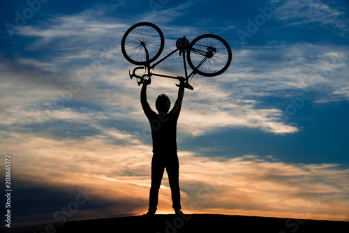 Man lifting bicycle at sunset sky. Silhouette of man lifts bicycle standing on hill on evening sky background. Enjoy your life.