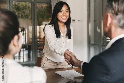 Business, career and placement concept - image from back of two employers sitting in office and shaking hand of young asian woman, after successful negotiations or interview