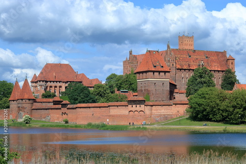 Zamek krzyżacki w Malborku, Polska, rzeka Nogat, zieleń wokół murów budowli, malownicze chmury na niebieskim niebie
