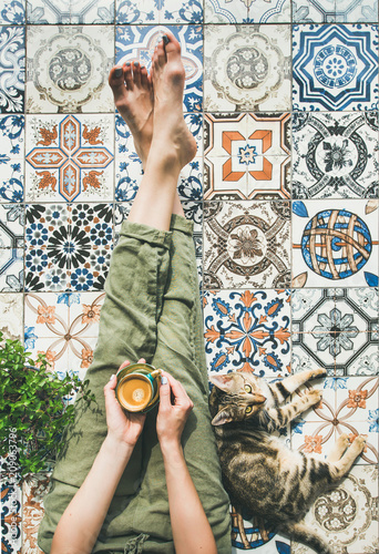 Lazy morning on terrace. Flat-lay of womans legs in cosy linen pants, plant, cat and cup of coffee in hand over colorful moroccan tile floor, top view