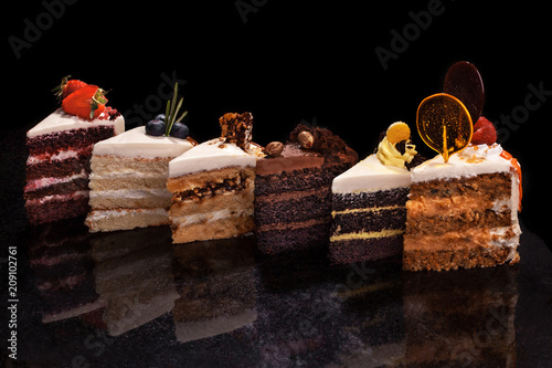 Assorted large pieces of different cakes: chocolate, raspberries, strawberries, nuts, blueberries. Pieces of cakes on a black table.