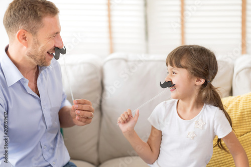 Happy father day. Dad and girl playing and having fun with mustache together at living room. Setup studio shooting.