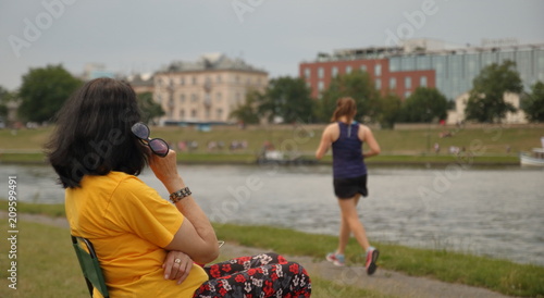 Czarnowłosa kobieta w żółtekj koszulce i kolorowych cienkich spodniach siedzi, bokiem, na nabrzeżu rzeki w mieście, trzyma okulary słoneczne, patrzy za uprawiającą jogging młodą kobietą w szortach
