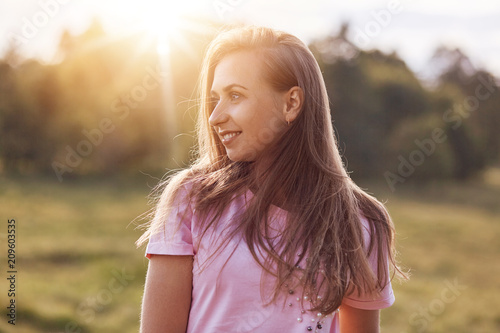 Pensive delighted happy young European female with long hair, looks thoughtfully aside, focused into distnace, dressed in casual t shirt, enjoys sunny summer day, dreams about something pleasant