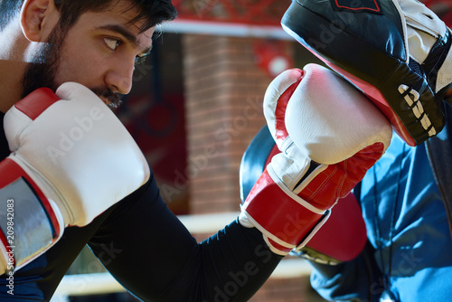 Side view of serious man in boxing gloves practicing with trainer wearing focus mitts. 