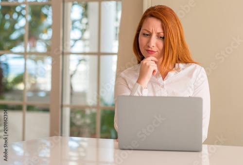 Redhead woman using computer laptop at home serious face thinking about question, very confused idea