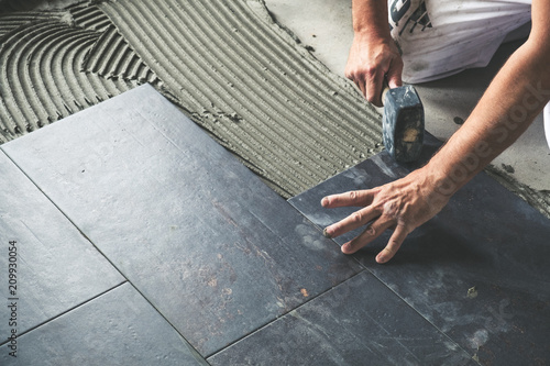 Worker placing ceramic floor tiles on adhesive surface, leveling with rubber hammer
