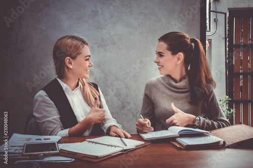 Two young european business women are sitting on the table and giving the thumbs up to agree on a decision about their work or project