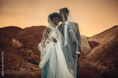 Newlyweds stand under bridal veil, smile and kiss in canyon at sunset.