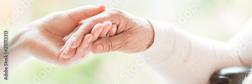 Close-up of tender gesture between two generations. Young woman holding hands with a senior lady. Blurred background. Panorama.