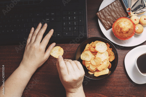 Unhealthy snack at workplace. Hands of woman working at computer and taking chips from the bowl. Bad habits, junk food, high calorie eating, weight gain and lifestyle concept